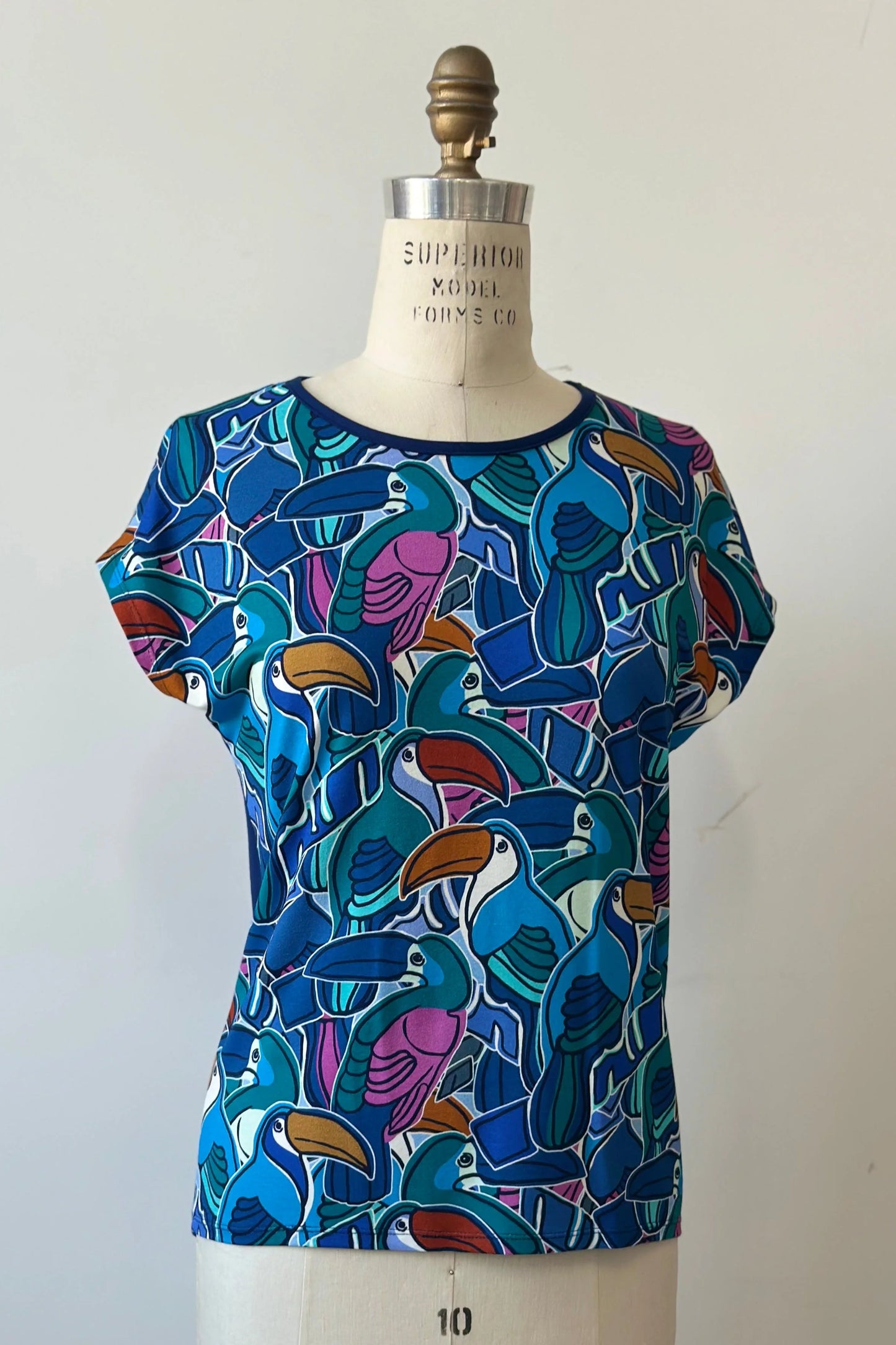 The Hanni Top by Kollontai in Admiral, a cap sleeved relaxed top with a jewel-toned parrot pattern on the front a solid blue back, is shown on a mannequin in front of a white background. 