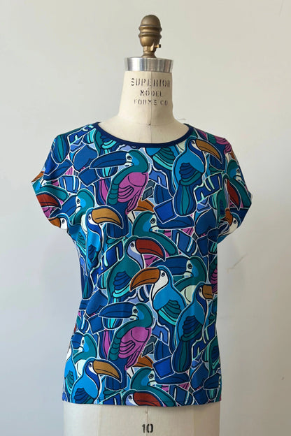 The Hanni Top by Kollontai in Admiral, a cap sleeved relaxed top with a jewel-toned parrot pattern on the front a solid blue back, is shown on a mannequin in front of a white background. 