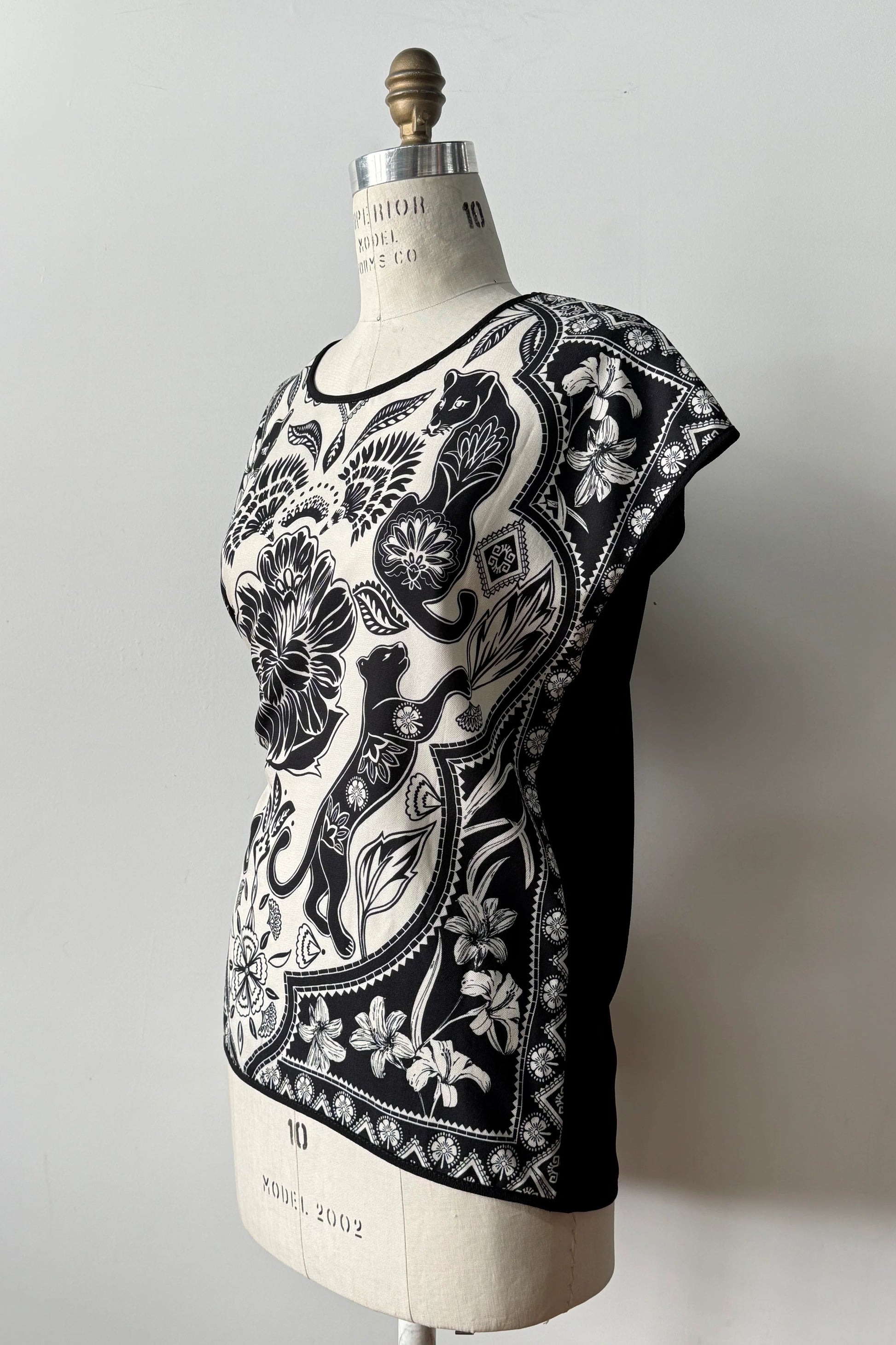 Side view of the Malo T-shirt from Kollontai, featuring a floral and panther print in black-on-ivory on the front, cap sleeves, and a solid black back, shown on a mannequin in front of a grey background 
