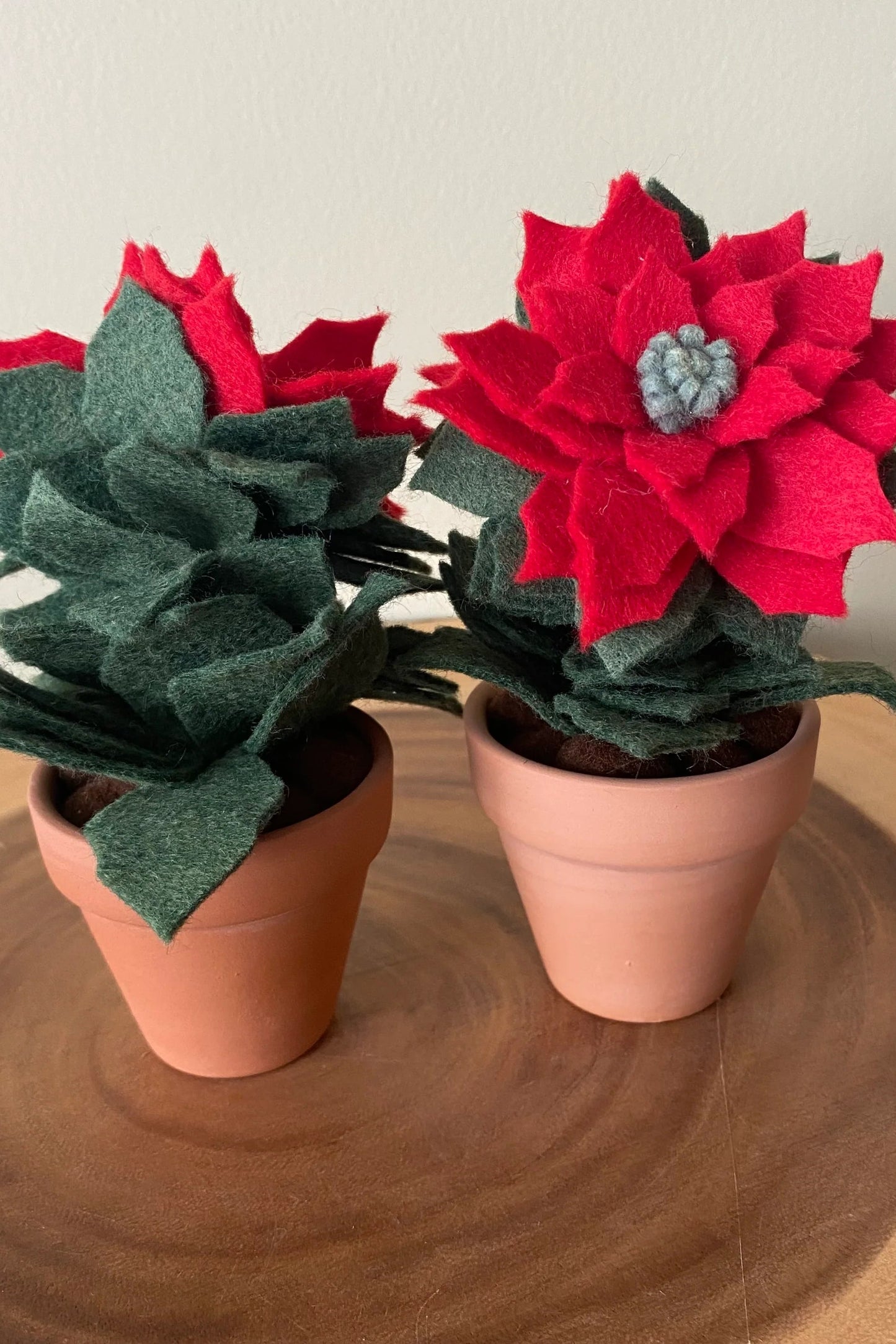 The Holiday Felt Poinsettia by ForgetBKnot is shown on a log against a white background. 