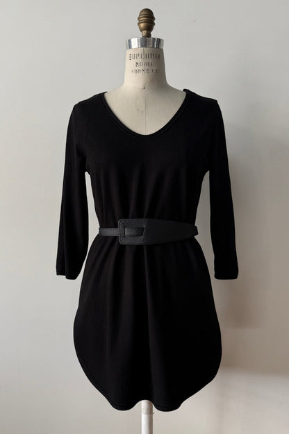 The Clara Tunic by Kollontai in Black, a stretch corduroy piece with a V-neck, a rounded hem, 3/4 sleeves, and inverted pleats at the shoulders, is shown on a mannequin against a grey background. 