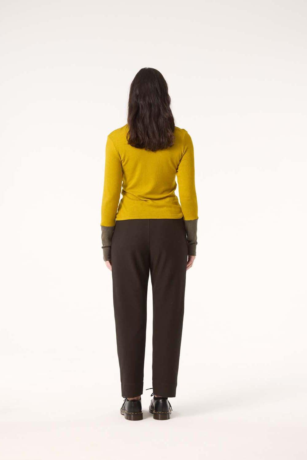 Back view of a woman wearing the Belief Pants by Cokluch in Pine with a yellow top, standing in front of a white background.