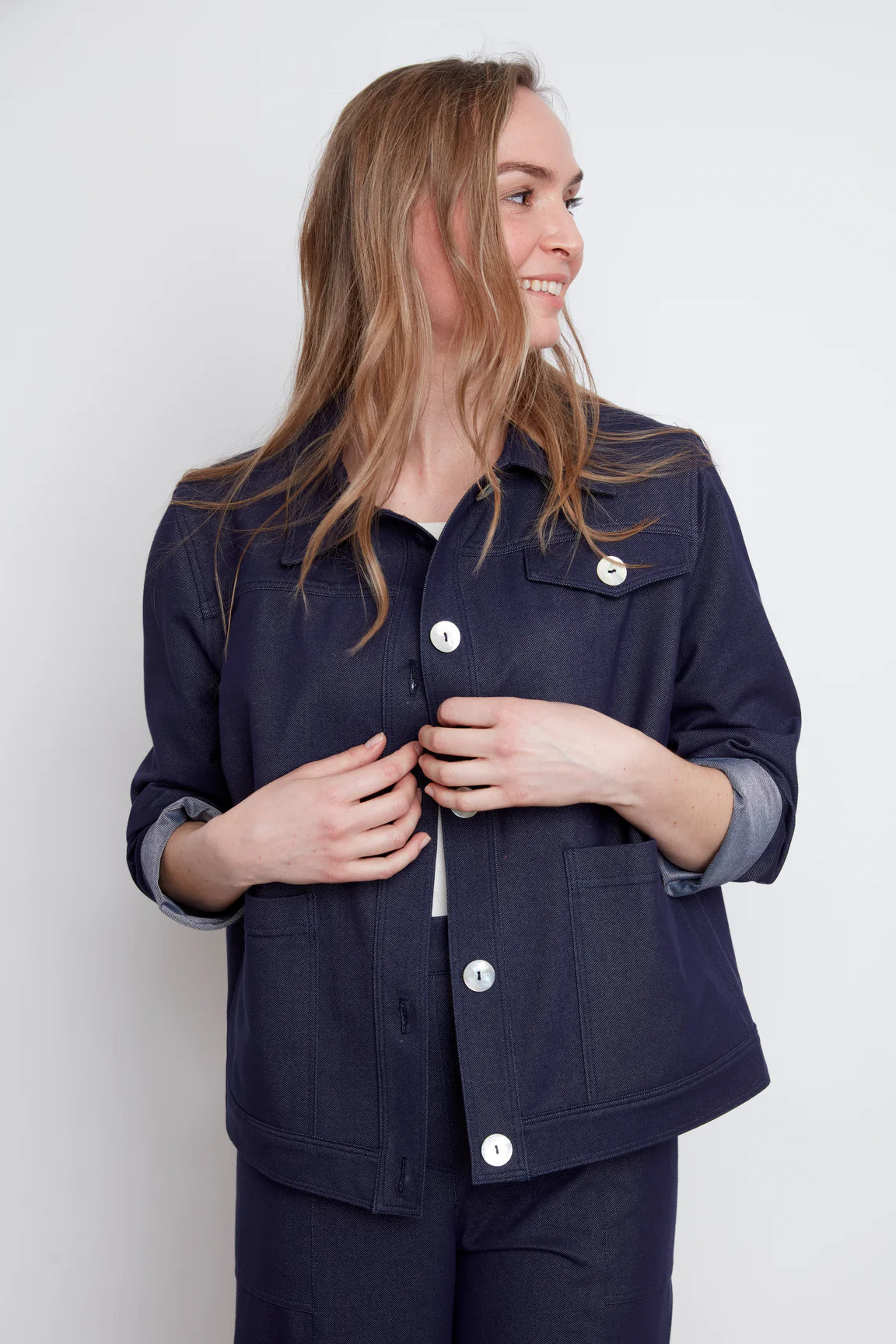 A woman wearing the Bella Knit Jacket by Julei, a denim style jacket with large contrast buttons, a chest pocket and two front patch pockets. She is wearing it with a white and the matching Brooke Knit Trousers and standing in front of a white wall. 