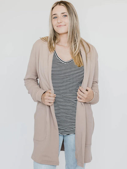 A woman wearing the Blondie Classic Briton Cardigan in Stucco, a long open front cardigan with large patch pockets. She is wearing it with jeans and a striped t-shirt and standing in front of a white background. 