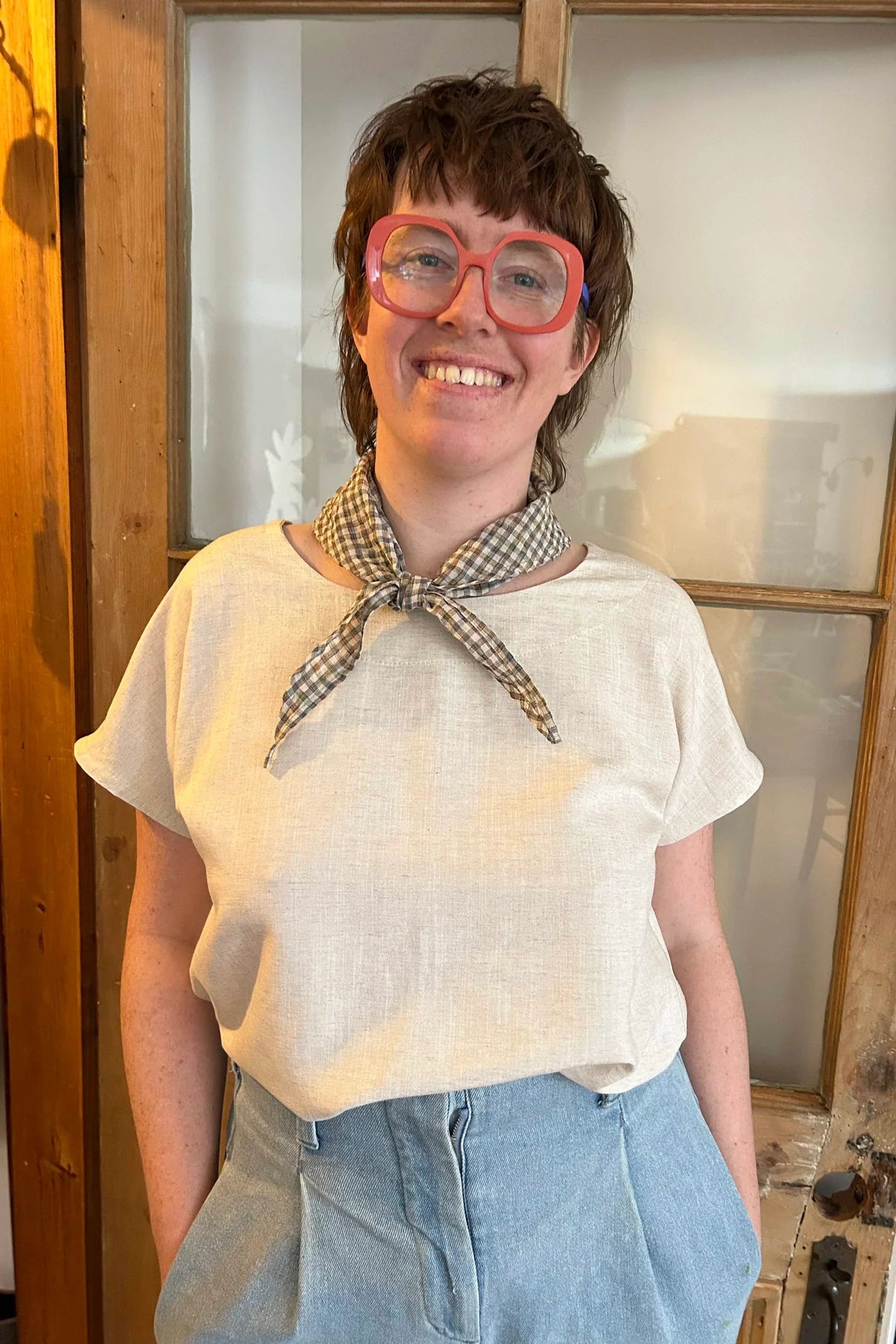 A woman wearing the Boronia Top by Kazak in Natural Linen with a round neck, short sleeves, slightly cropped and boxy fit, with a box pleat at the back yoke. She is wearing it with a checkered scarf and light jeans and standing in front of a wooden door. 