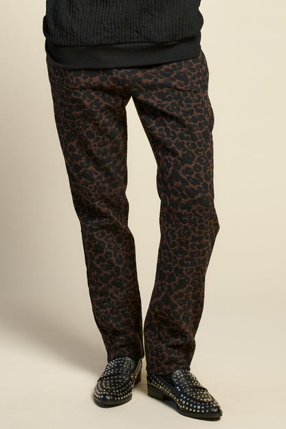 Waist down view of a woman wearing the Breeze pants from Cokluch in Cappuccino Leopard, featuring a slim fit, an attached belt with a D-ring closure, and pockets, all in a chic ankle-length style. She is wearing them with a black top and standing in front of a beige background. 