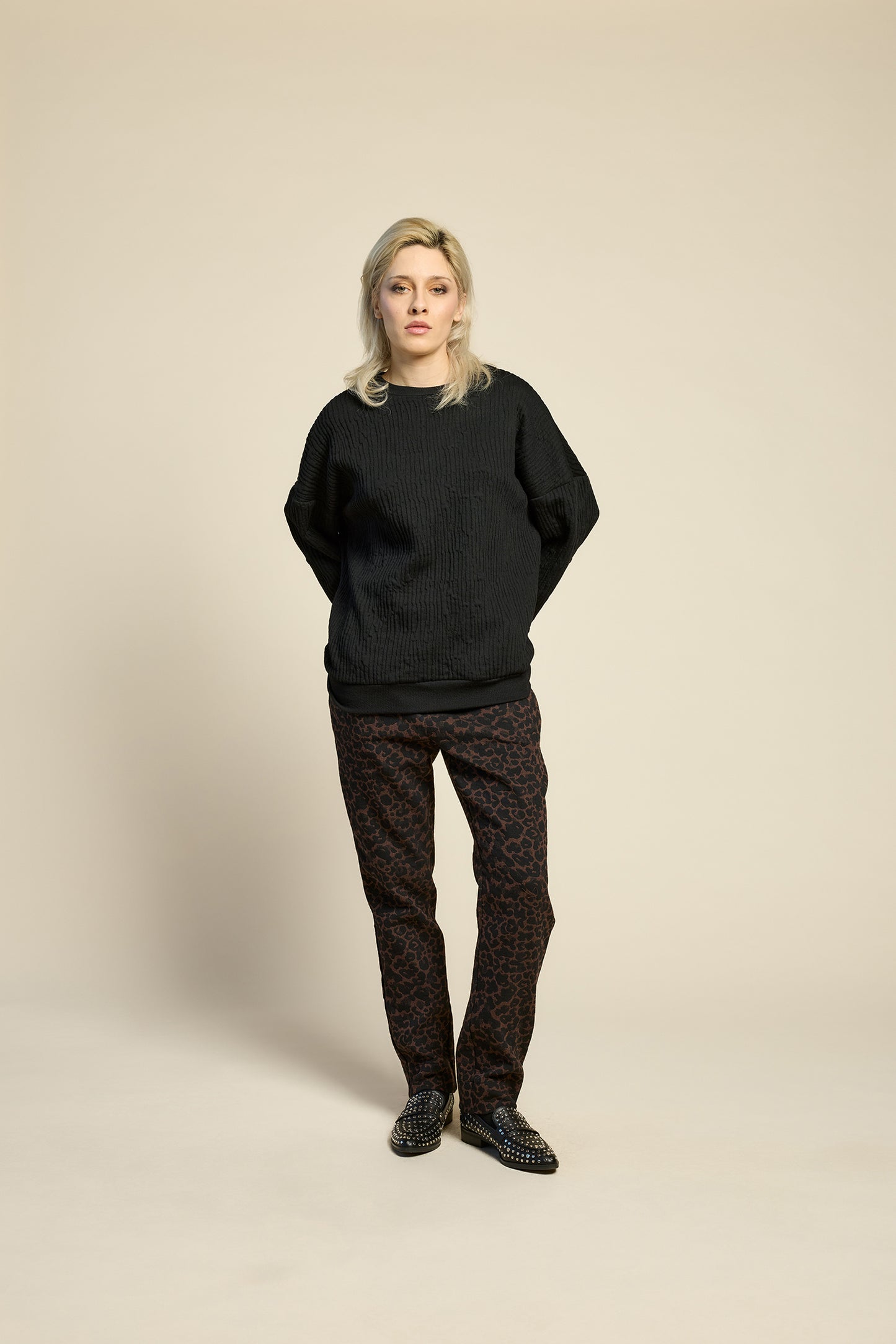A woman wearing the Breeze pants from Cokluch in Cappuccino Leopard, featuring a slim fit, an attached belt with a D-ring closure, and pockets, all in a chic ankle-length style. She is wearing them with a black top and standing in front of a beige background. 
