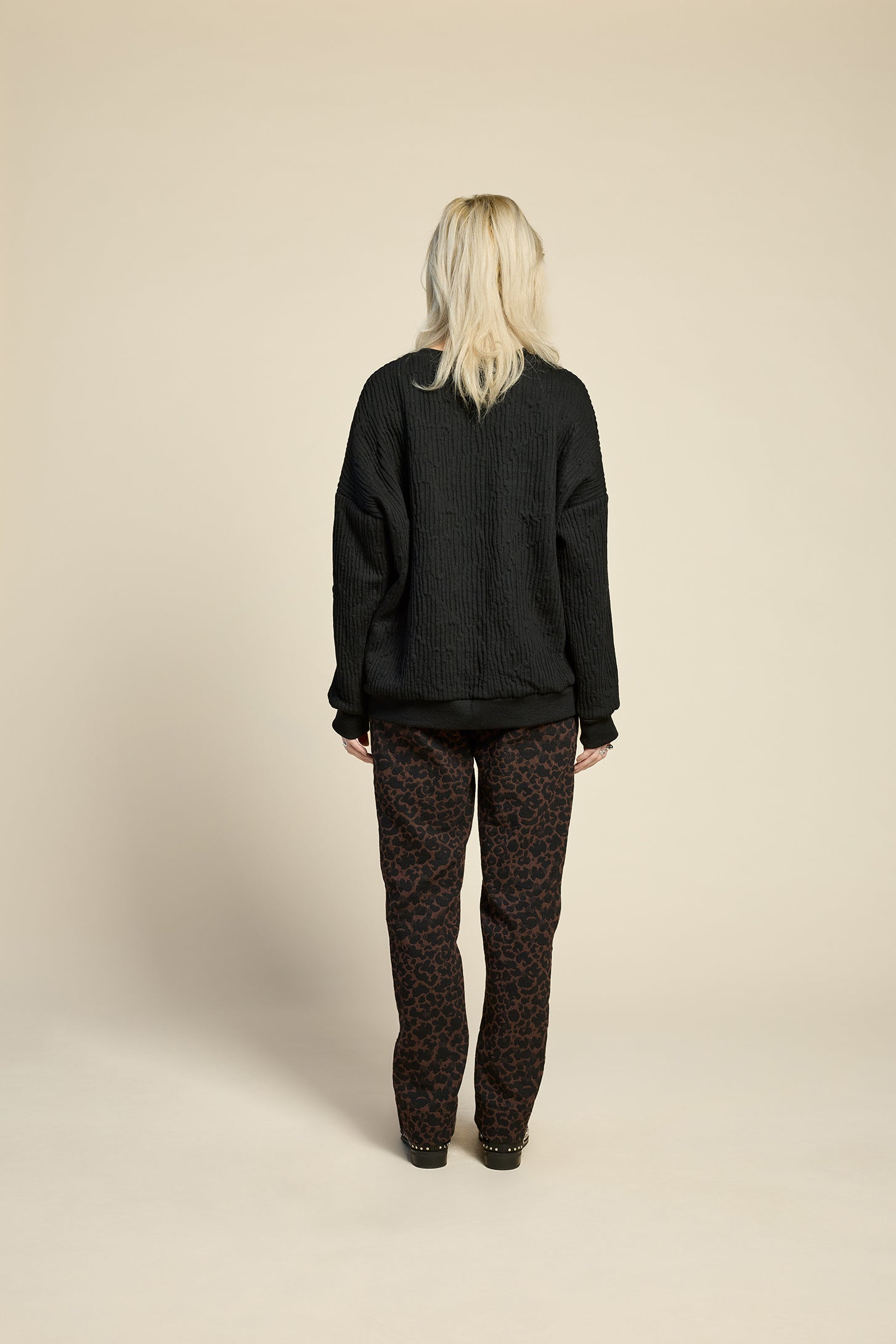 Back view of a woman wearing the Breeze pants from Cokluch in Cappuccino Leopard, featuring a slim fit, an attached belt with a D-ring closure, and pockets, all in a chic ankle-length style. She is wearing them with a black top and standing in front of a beige background. 