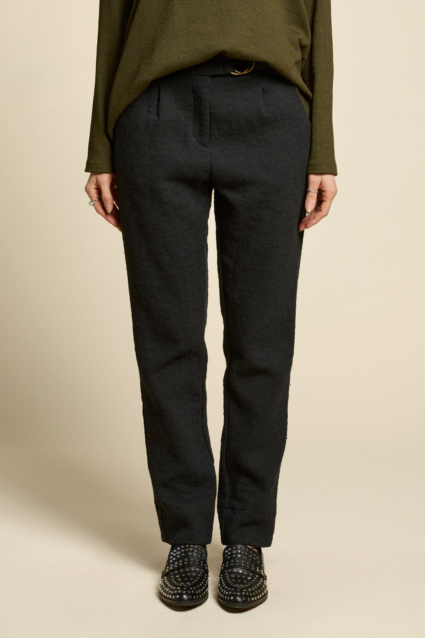 Waist down view of a woman wearing the Breeze pants from Cokluch in textured Black fabric, featuring a slim fit, an attached belt with a D-ring closure, and pockets, all in a chic ankle-length style. She is wearing them with a black top and standing in front of a beige background. 