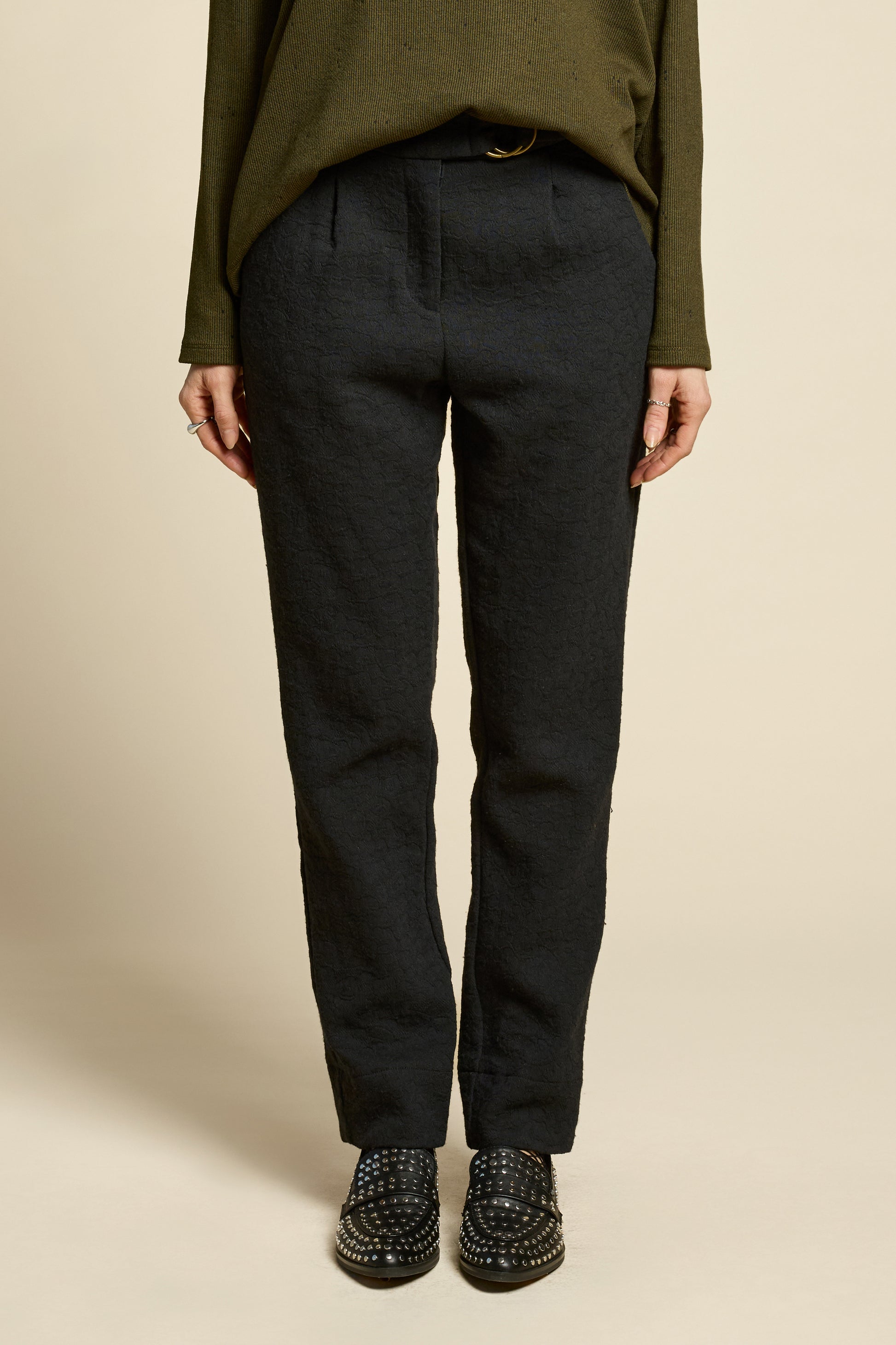 Waist down view of a woman wearing the Breeze pants from Cokluch in textured Black fabric, featuring a slim fit, an attached belt with a D-ring closure, and pockets, all in a chic ankle-length style. She is wearing them with a black top and standing in front of a beige background. 
