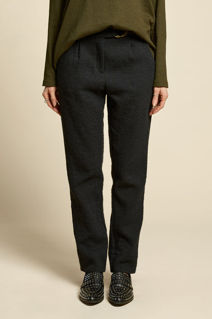 Waist down view of a woman wearing the Breeze pants from Cokluch in textured Black fabric, featuring a slim fit, an attached belt with a D-ring closure, and pockets, all in a chic ankle-length style. She is wearing them with a black top and standing in front of a beige background. 