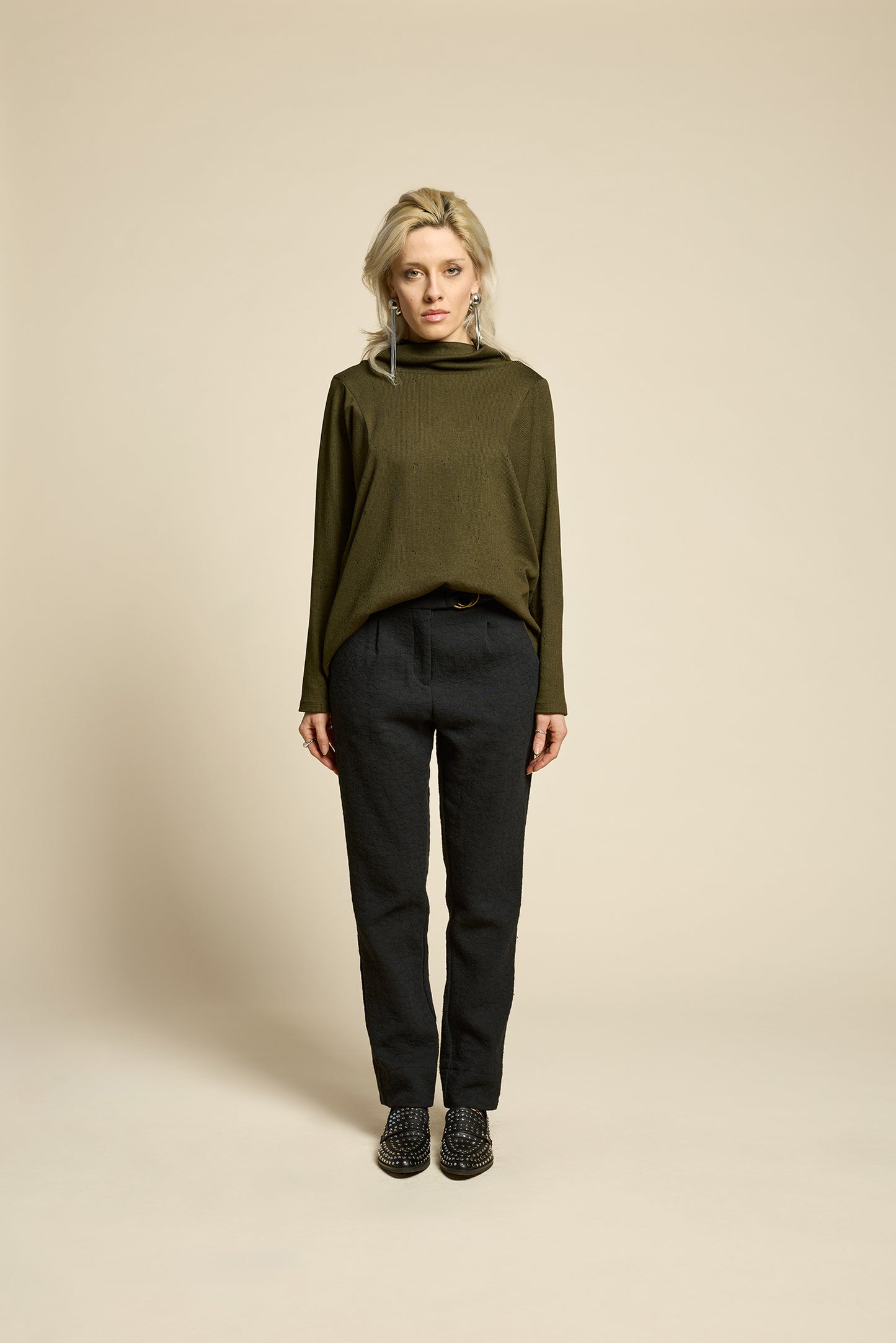 A woman wearing the Breeze pants from Cokluch in textured Black fabric, featuring a slim fit, an attached belt with a D-ring closure, and pockets, all in a chic ankle-length style. She is wearing them with a green top and standing in front of a beige background. 