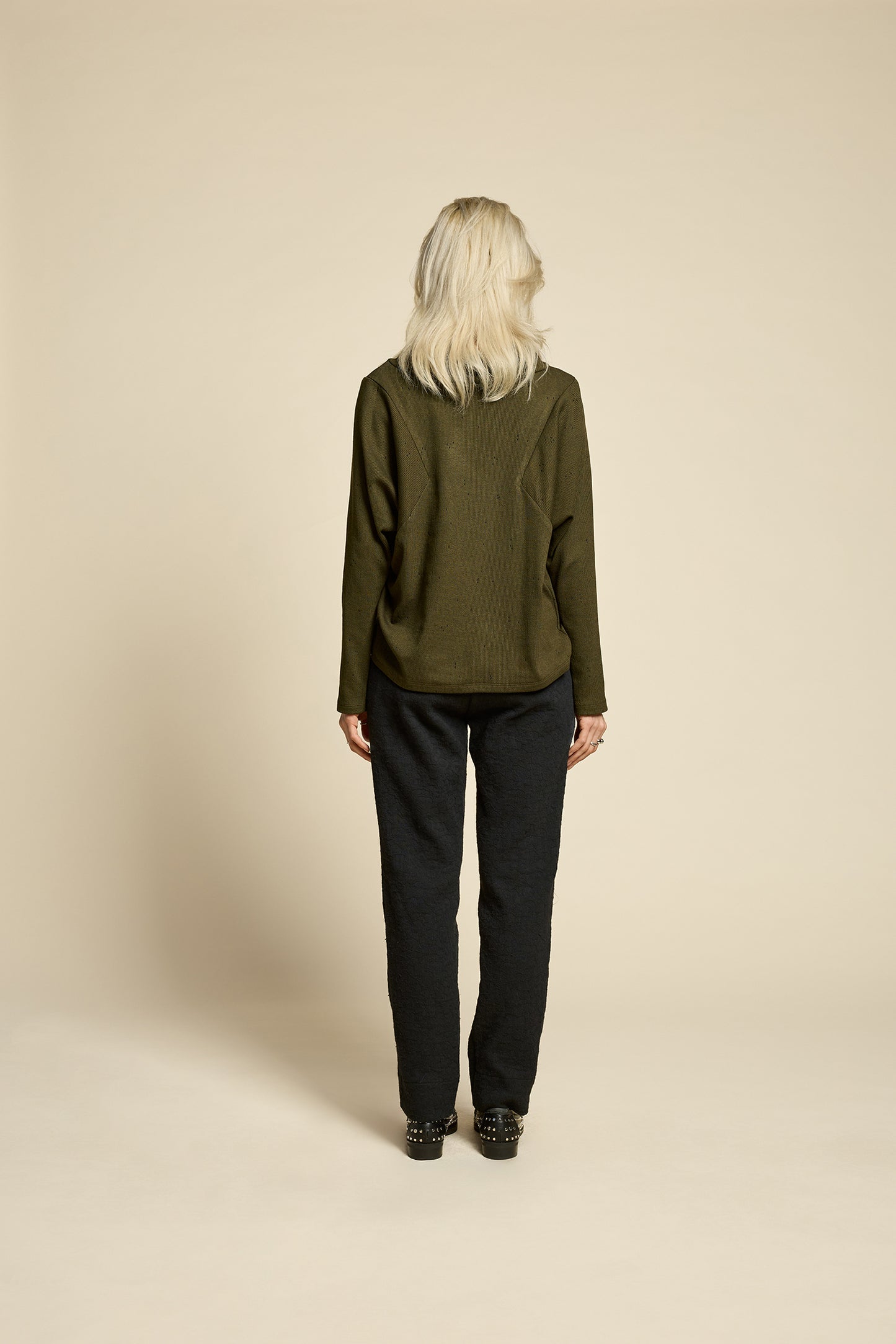 A woman wearing the Breeze pants from Cokluch in textured Black fabric, featuring a slim fit, an attached belt with a D-ring closure, and pockets, all in a chic ankle-length style. She is wearing them with a green top and standing in front of a beige background. 