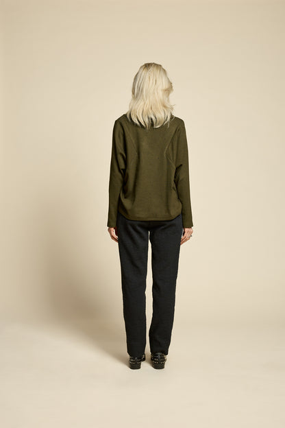 A woman wearing the Breeze pants from Cokluch in textured Black fabric, featuring a slim fit, an attached belt with a D-ring closure, and pockets, all in a chic ankle-length style. She is wearing them with a green top and standing in front of a beige background. 