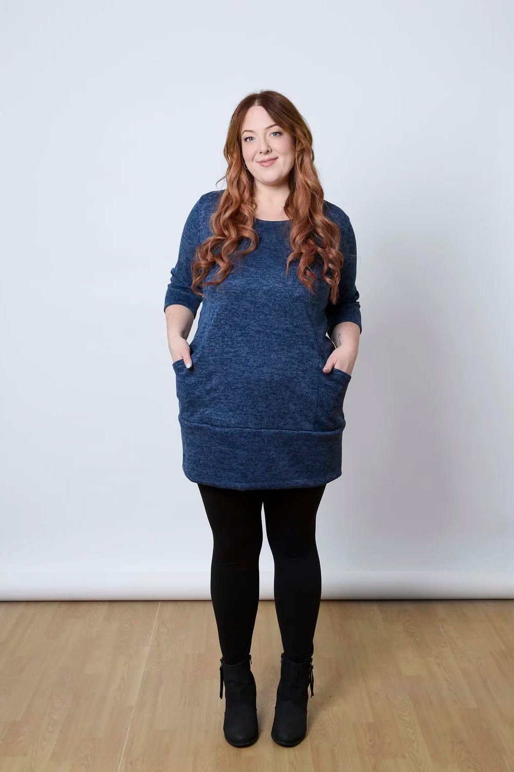 A woman wearing the Brooke Tunic by Tangente in Indigo, with 3/4 sleeves and a fitted band at the bottom. She is wearing it with black leggings and standing against a white wall. 