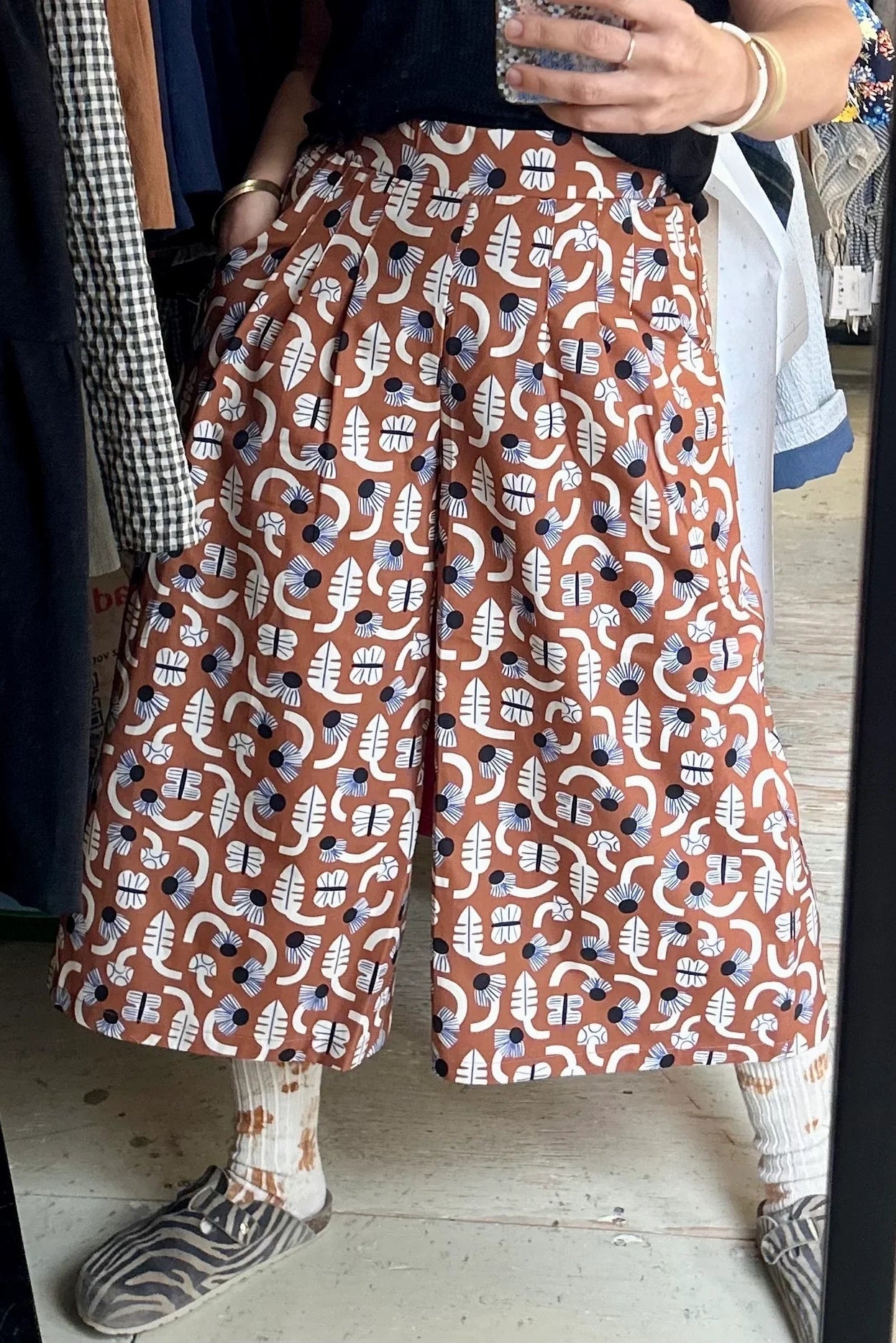 Waist down view of a woman wearing the Petunia Pants by Kazak in Foraging, wide legged cropped pants with a waistband that's smooth in front but elastic at the back, front pleats, and pockets. She is wearing them with a black top and standing amidst clothing racks.