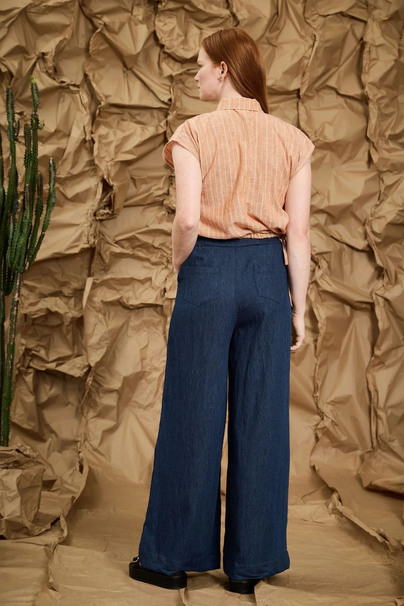Back view of a woman wearing the Matisse Trousers by Melow in Denim, full length wide leg pants with a zipper front and a high waist with elastic at the back with side and back patch pockets. She is wearing them with a peach top and standing in front of a textured brown backdrop