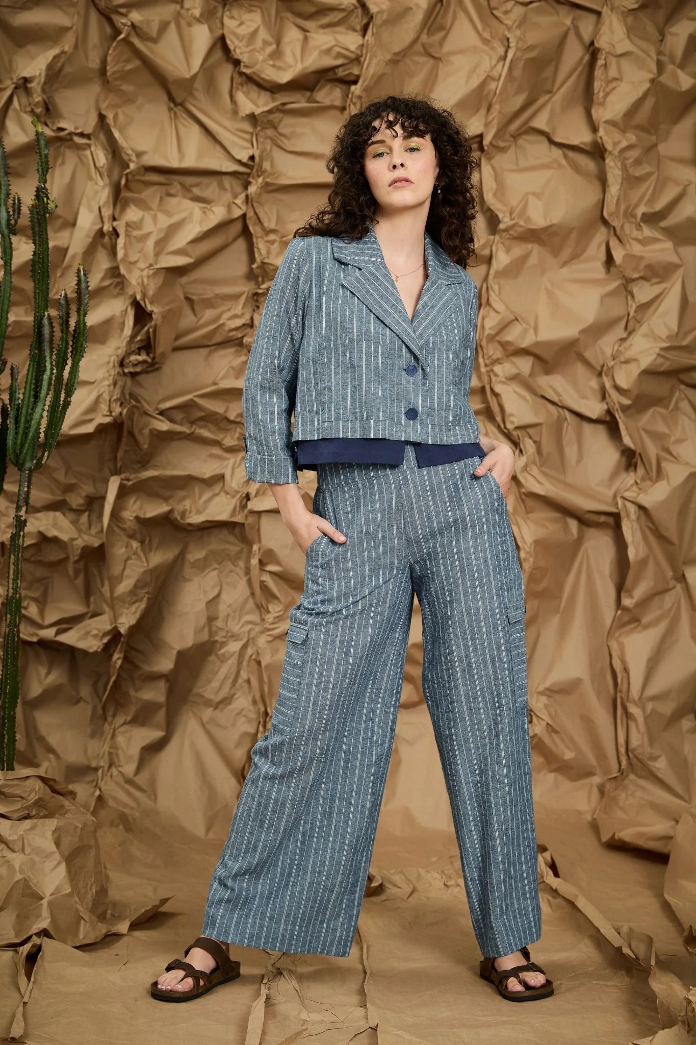 A woman wearing the Maxime Pants by Melow in Blue stripe, straight legged pants with cargo flap pockets on the legs, a high wide waistband, and an invisible side zipper. She is wearing them with a matching Marou Jacket and standing against a textured brown background