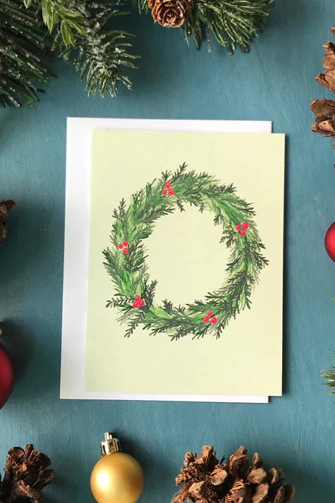 A card from a box set by Carabara Designs, featuring a Winter Wreath design on a pale green background, is shown against a teal background with holiday decor around it. 