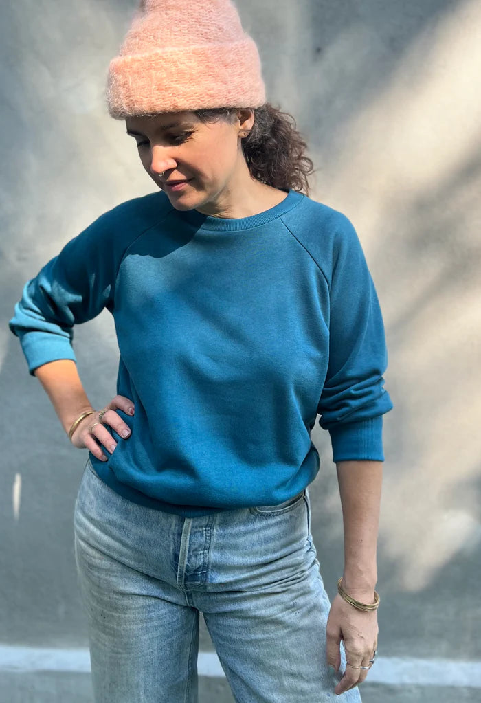 A woman wearing the Lipa Crewneck Sweatshirt by Kazak in Teal, featuring a round neck, 3/4 raglan sleeves, and a fitted band across the bottom. She is wearing it with jeans and a pink tuque and is standing outside in front of a grey wall.