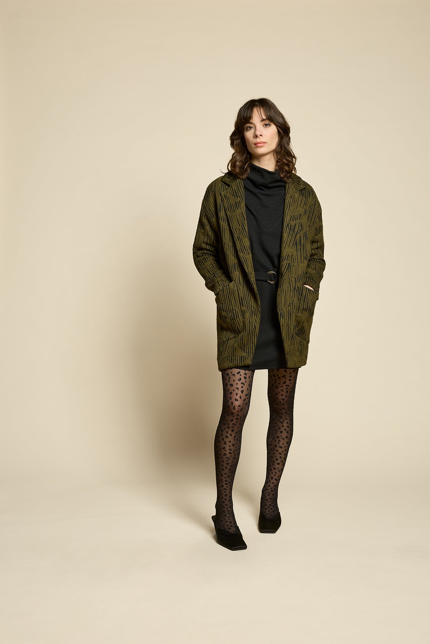 A woman wearing the Call Me Jacket by Cokluch in Olive, a thigh-length open collared jacket in unique Crinkle Max fabric with two large patch pockets. She is wearing it over a black outfit on a beige background.