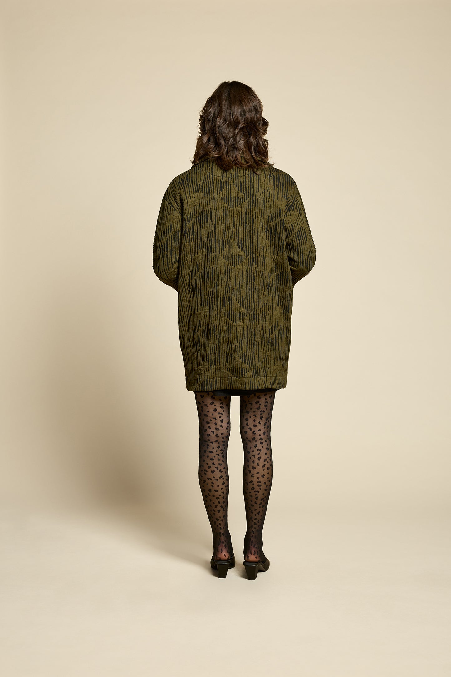 Back view of a woman wearing the Call Me Jacket by Cokluch in Olive, a thigh-length open collared jacket in unique Crinkle Max fabric with two large patch pockets. She is wearing it over a black outfit on a beige background.