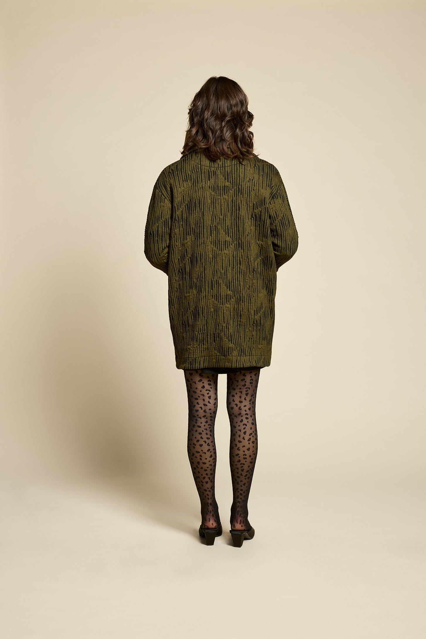 Back view of a woman wearing the Call Me Jacket by Cokluch in Olive, a thigh-length open collared jacket in unique Crinkle Max fabric with two large patch pockets. She is wearing it over a black outfit on a beige background.