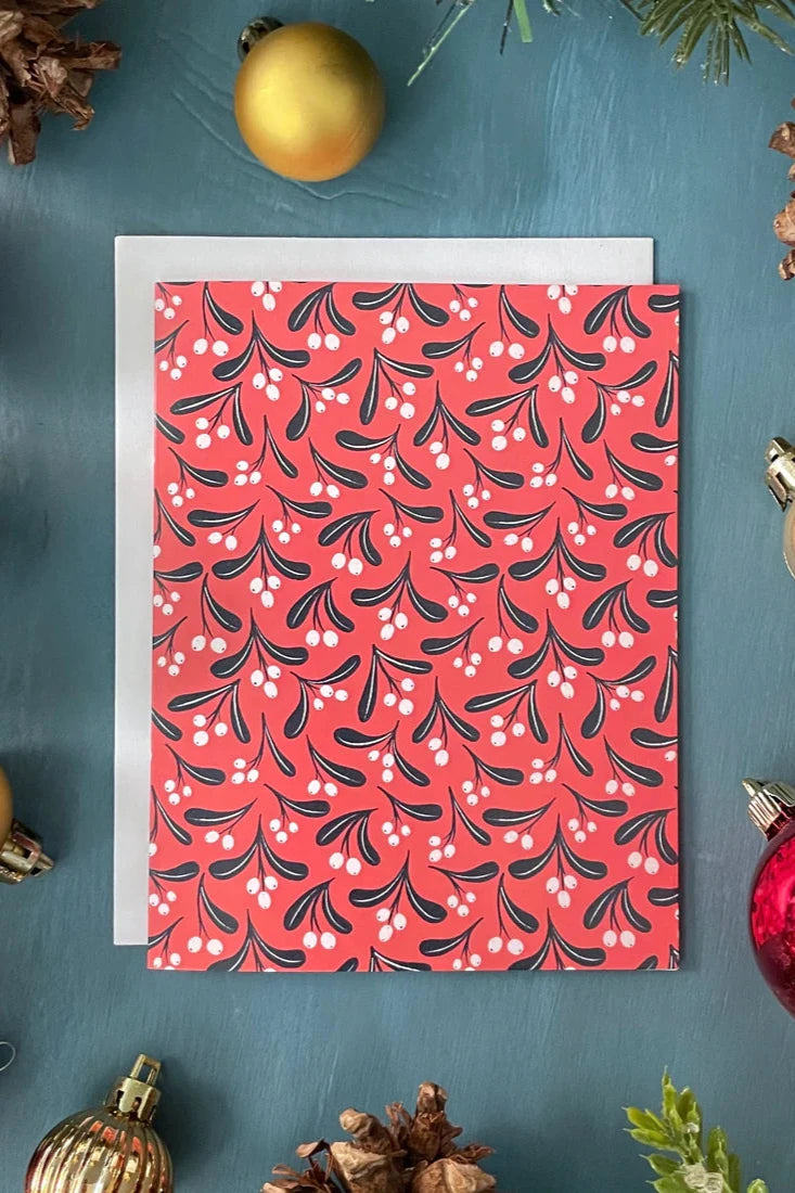 A Coastal Mistletoe card from a box set by Carabara Designs, featuring a mistletoe pattern on a red background, is shown against a teal background with holiday decor around it.