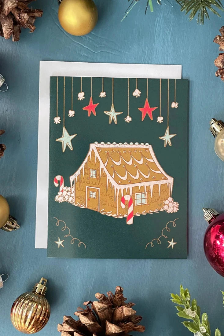 A card from a box set by Carabara Designs, featuring a Midcentury Gingerbread House design, is shown against a teal background with holiday decor around it. 