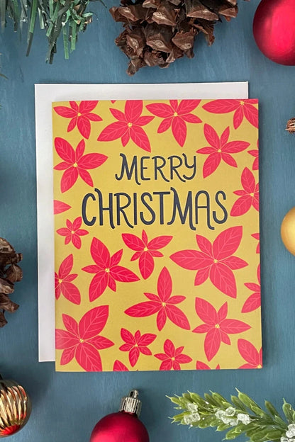 A card from a box set by Carabara Designs, featuring poinsettias on a yellow background and the words "Merry Christmas", is shown against a teal background with holiday decor around it. 