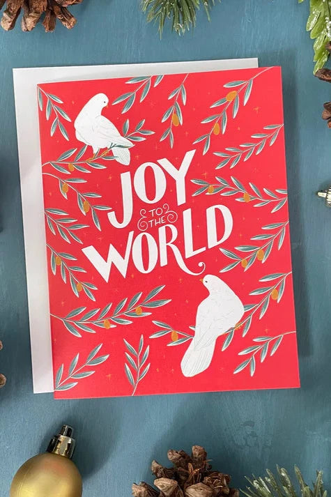 A card from a box set by Carabara Designs, featuring white doves and the words "Joy to the World" on a red background, is show against a teal background with holiday decor around it. 