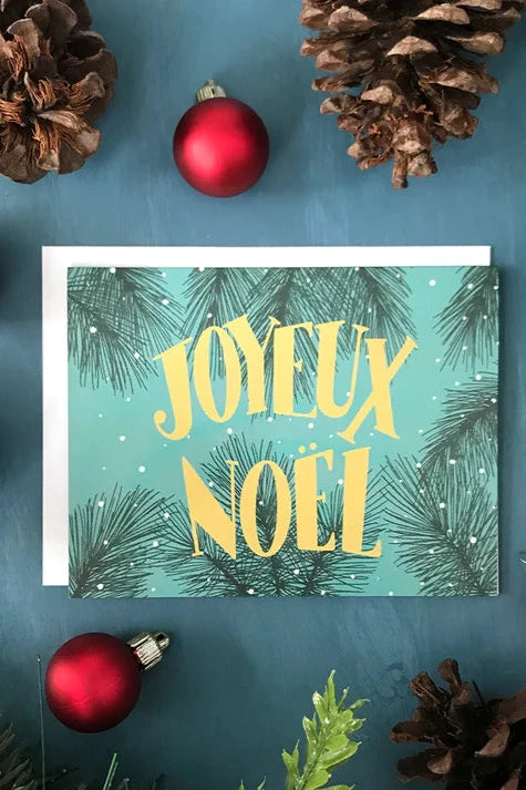 A card from a box set by Carabara Designs, featuring the words "Joyeux Noel" in yellow with evergreen needles in the background, is shown against a teal background with holiday decor around it. 