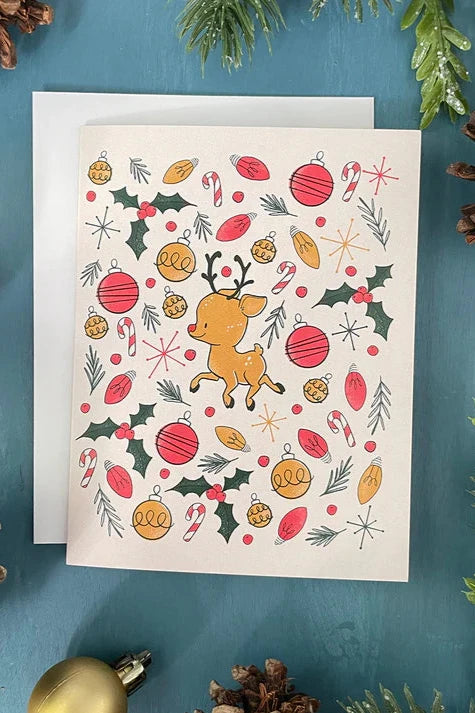 A card from a box set by Carabara Designs, featuring a Midcentury Reindeer pattern on a white  background, is shown against a teal background with holiday decor around it. 