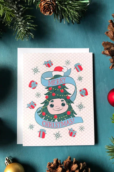 A card from a box set by Carabara Designs, featuring Nova Scotia's famed Woody the Christmas Tree, is shown against a teal background with holiday decor around it. 