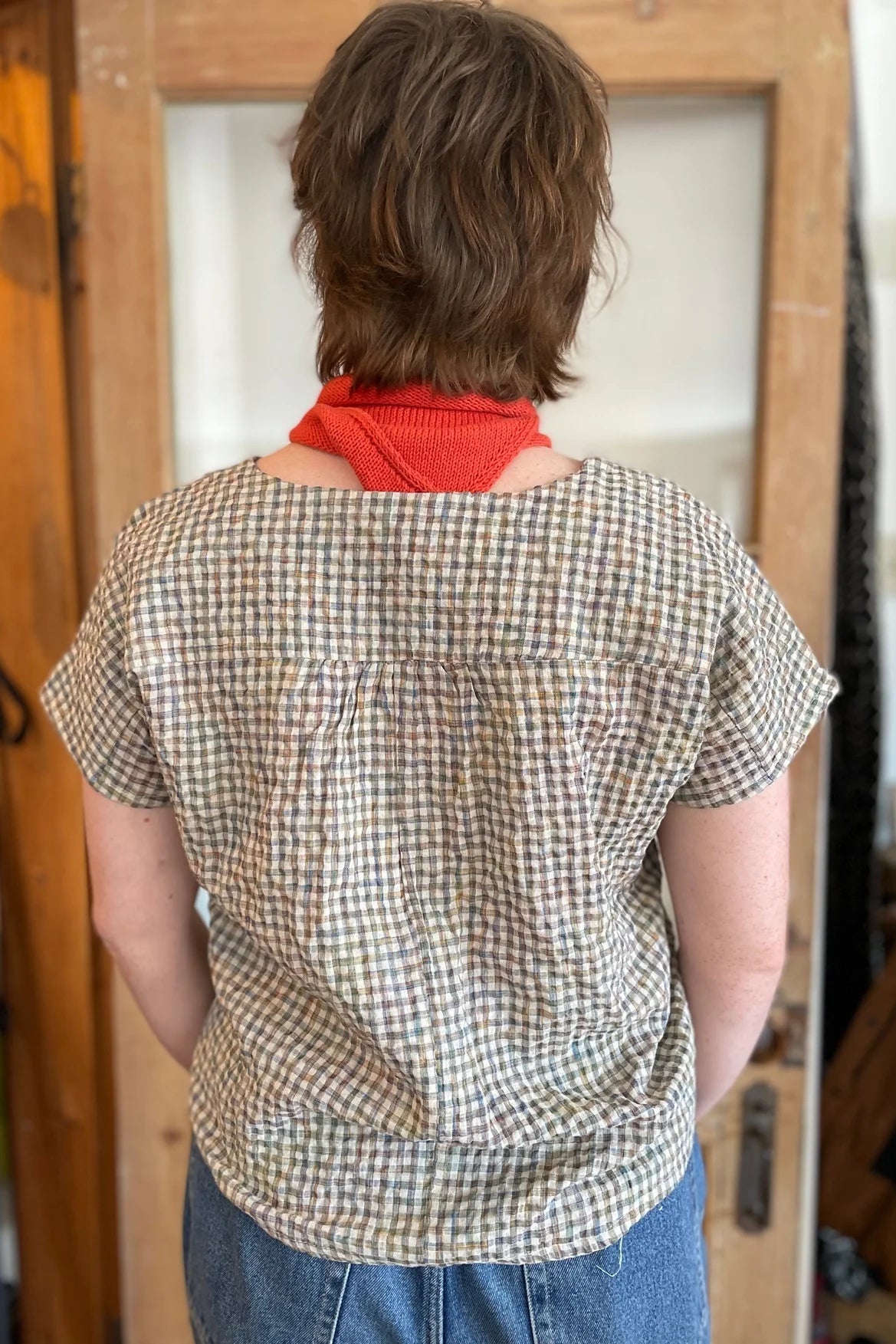 Back view of a woman wearing the Boronia Top by Kazak in Multicoloured Tiles with a round neck, short sleeves, slightly cropped and boxy fit, witha box pleat at the back yoke. She is wearing it with a red scarf and light jeans and standing in front of a wooden door. 