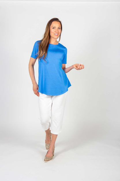 A woman wearing the Catherine Tee by Pure Essence in Mykonos Blue with white pants, standing in front of a white background