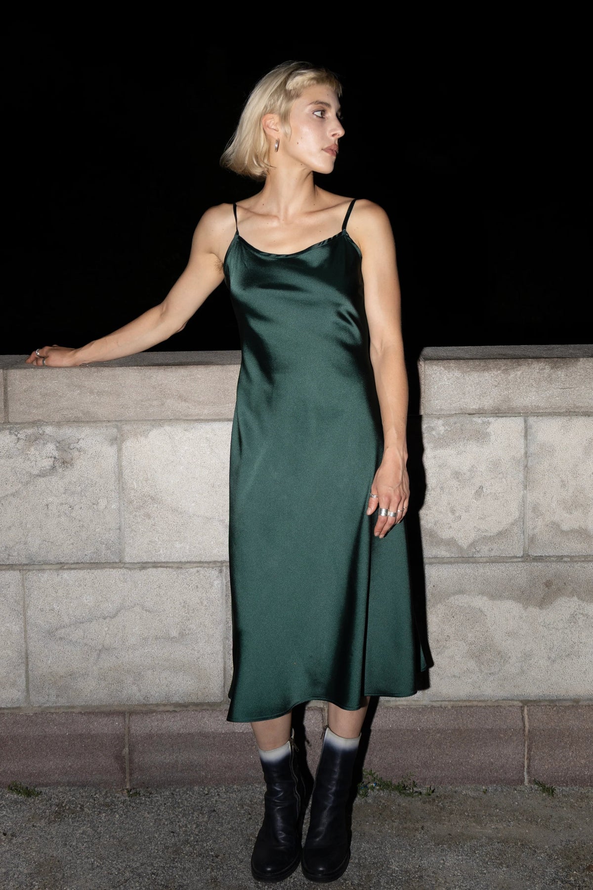 A woman wearing the Celestite Dress by Eve Lavoie in Green Satin, a slip dress with slanted spaghetti straps and an opening at the back with a tie detail. It falls to mid calf. She is standing outdoors by a stone wall. 