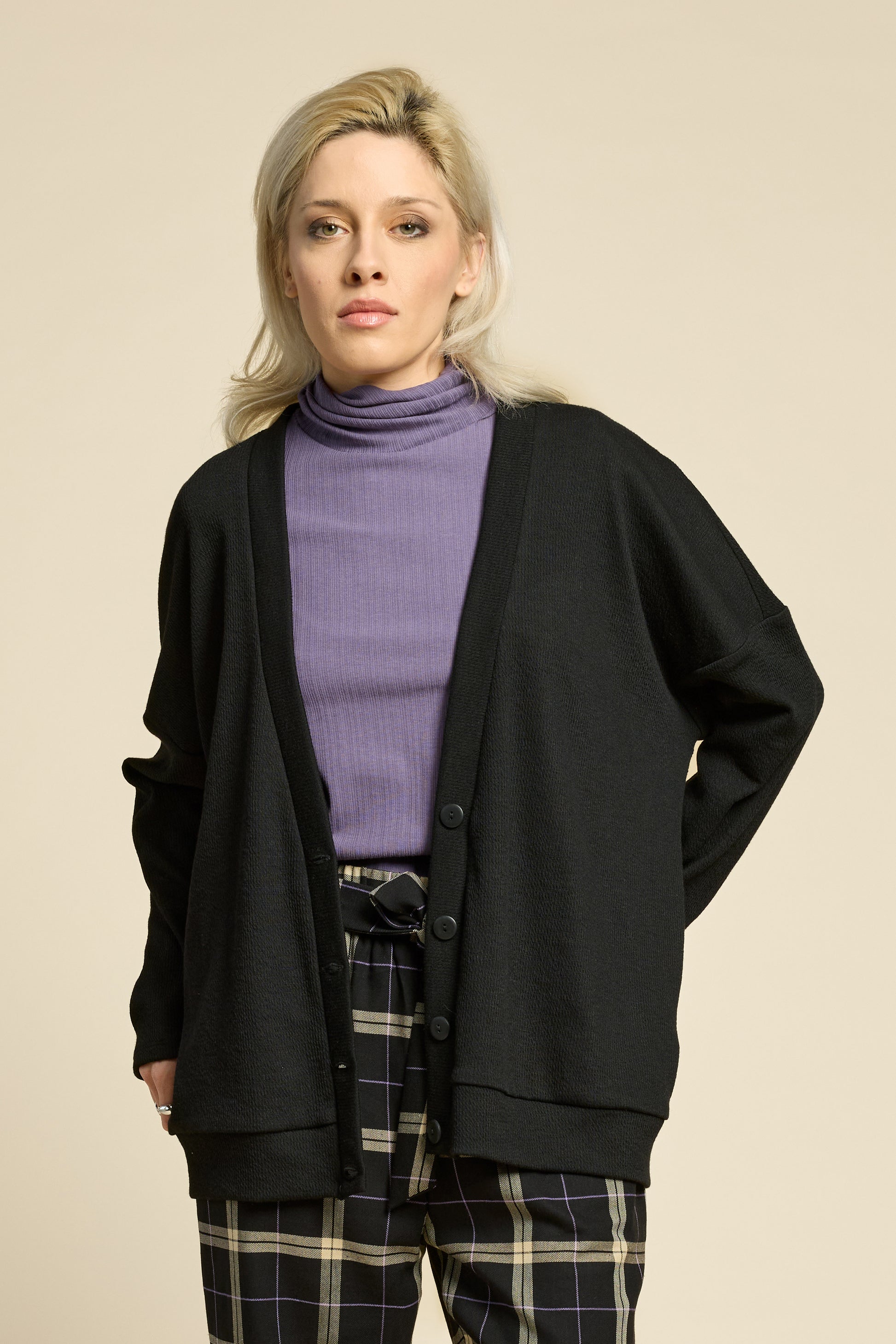Waist up view of a woman wearing the Cendres Cardigan by Cokluch in Black, a slightly oversized cardigan in dotted fabric with four buttons. She is wearing it with a purple top and the West Coast pants in Black Plaid and standing in front of a beige background. 