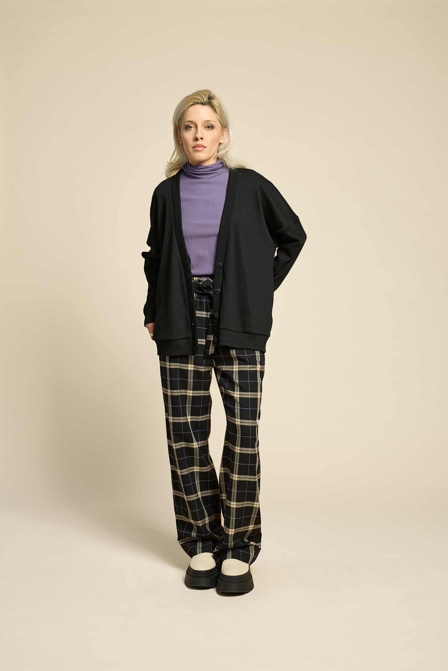 Waist up view of a woman wearing the Cendres Cardigan by Cokluch in Black, a slightly oversized cardigan in dotted fabric with four buttons. She is wearing it with a purple top and the West Coast pants in Black Plaid and standing in front of a beige background. 
