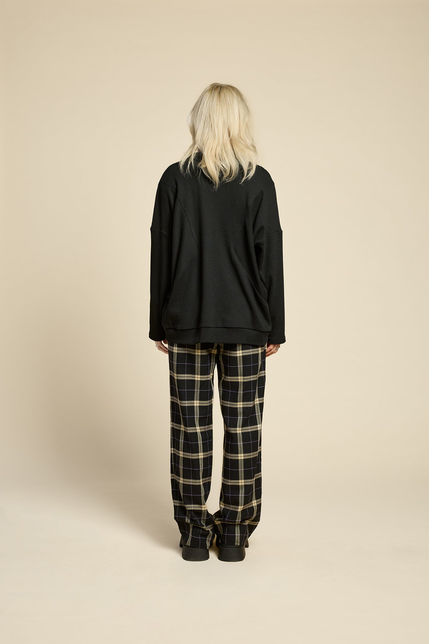 Back view of a woman wearing the Cendres Cardigan by Cokluch in Black, a slightly oversized cardigan in dotted fabric with four buttons. She is wearing it with a purple top and the West Coast pants in Black Plaid and standing in front of a beige background. 