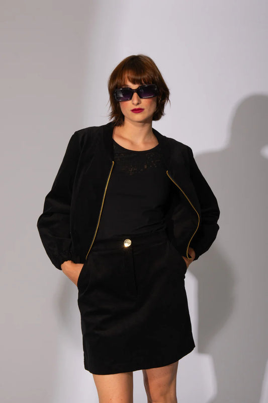 A woman wearing the Burning Miniskirt by Eve Lavoie in Black, a short skirt with a front zip and a large gold button, side slash pockets, and a slit at the back. she is wearing it with a black jacket and standing in front of a white background. 