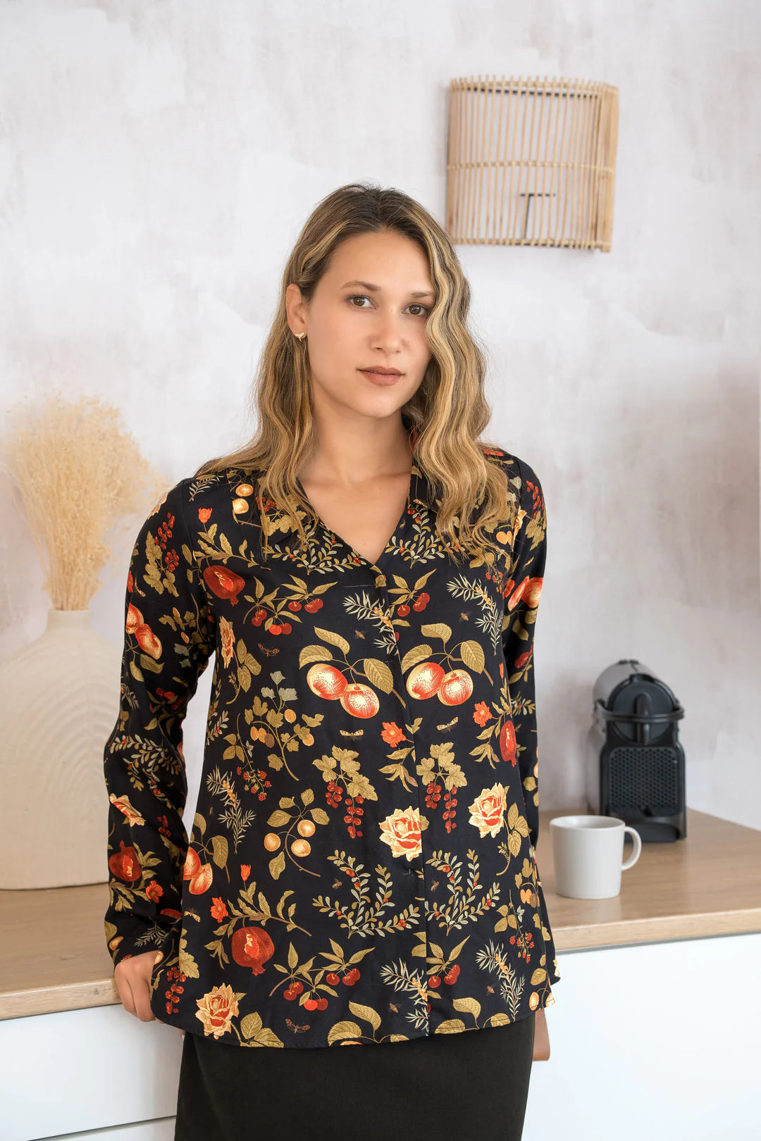 A woman wearing the Martini Blouse by Cherry Bobin in Pomegranate Apples print, a button front shirt with a classic collar. She is wearing it with black pants and leaning against a counter. 
