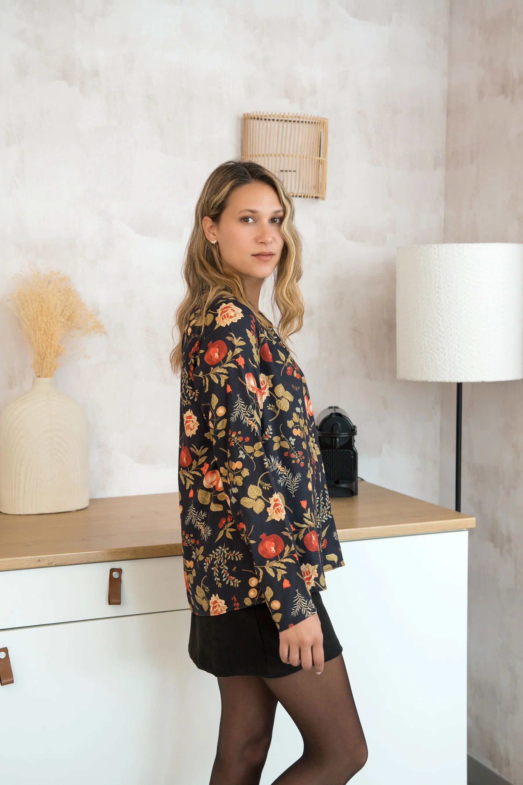 Side view of a woman wearing the Martini Blouse by Cherry Bobin in Pomegranate Apples print, a button front shirt with a classic collar. She is wearing it with black skirt and standing by a counter. 