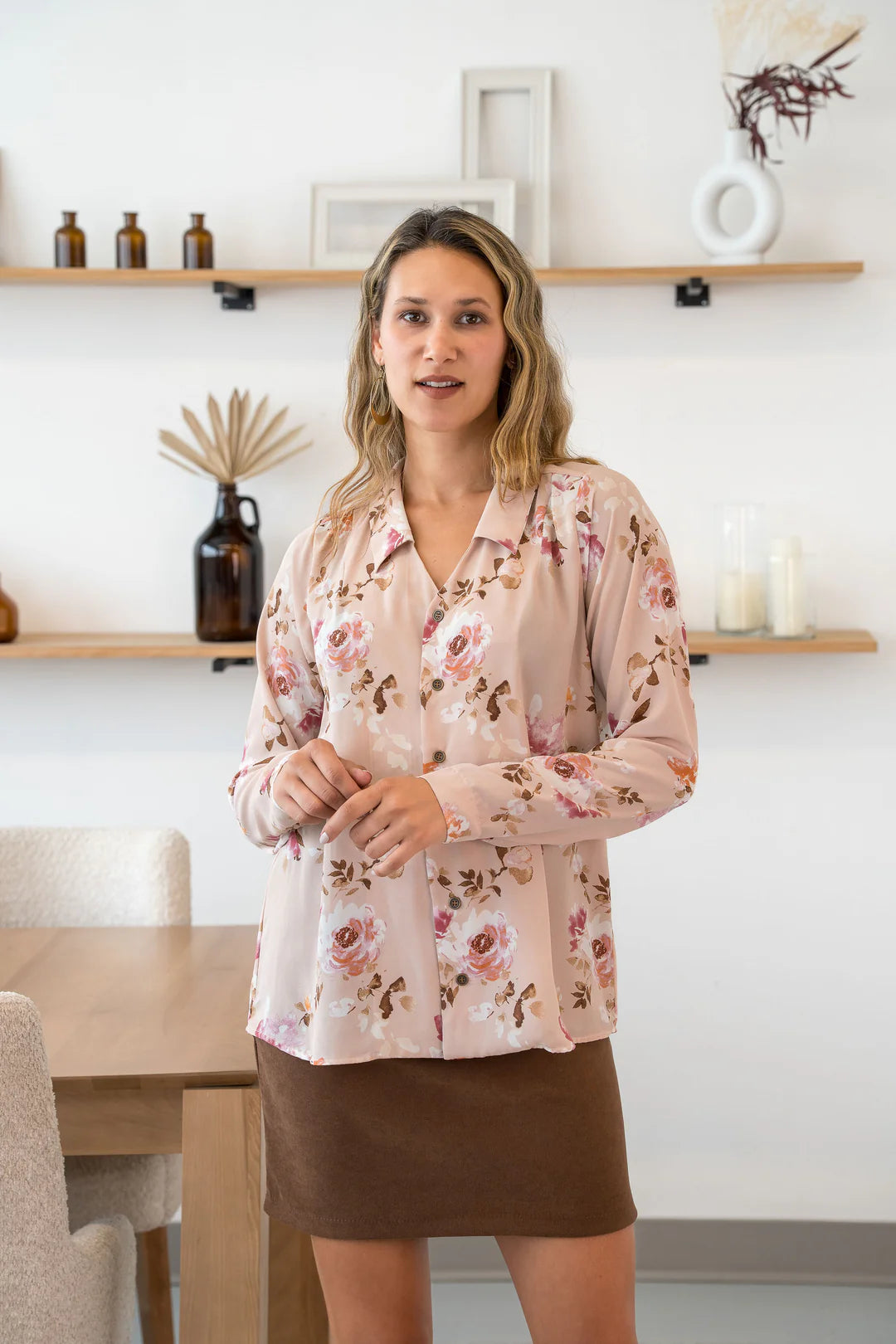 A woman wearing the Martini Blouse by Cherry Bobin in pale pink Floral, a button front shirt with a classic collar. She is wearing it with a brown skirt and standing by a table. 