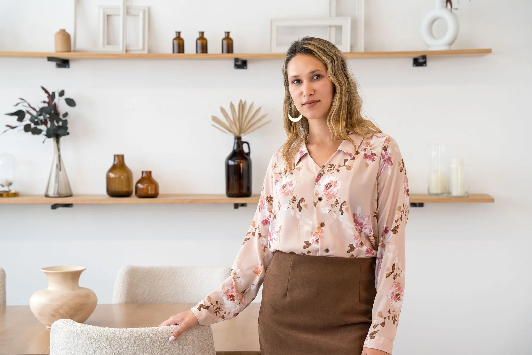 A woman wearing the Martini Blouse by Cherry Bobin in pale pink Floral, a button front shirt with a classic collar. She is wearing it with a brown skirt and standing by a table. 