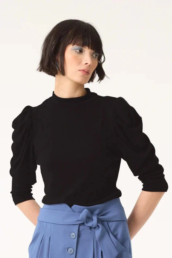 Semi close-up of woman wearing the Nenuphar Top by Cokluch in Black Bamboo, featuring a high collar and 3/4 length puffed sleeves, with light blue pants. She standing in front of a white background. 