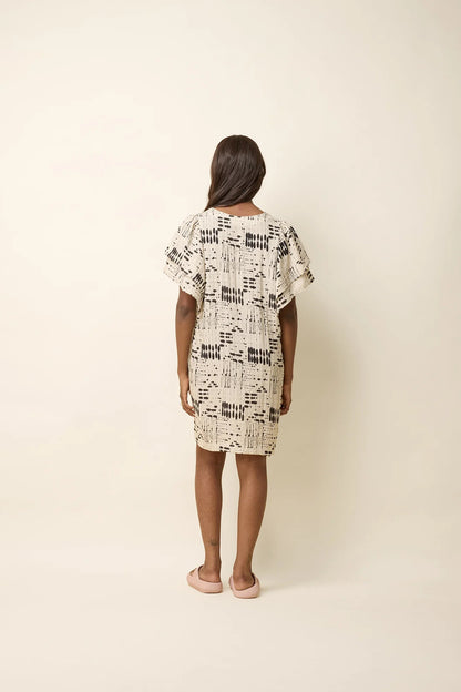 A woman wearing the Paramida Dress by Cokluch in Natural Ink, featuring a v-neck, a double ruffle on the short sleeves, and a loose fit that falls above the knee. She is standing in front of a beige background. 