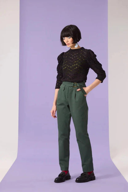 A woman wearing the Saules Pants by Cokluch in Eden, with a fly closure, an attached D-ring belt, a button and tab detail, side pockets, and an ankle length, made from OEKO-TEX certified fabric. She is wearing them with a black top and standing in front of a purple background. 