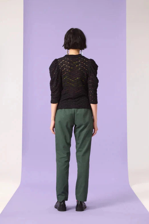 Back view of a woman wearing the Saules Pants by Cokluch in Eden, with a fly closure, an attached D-ring belt, a button and tab detail, side pockets, and an ankle length, made from OEKO-TEX certified fabric. She is wearing them with a black top and standing in front of a purple background. 