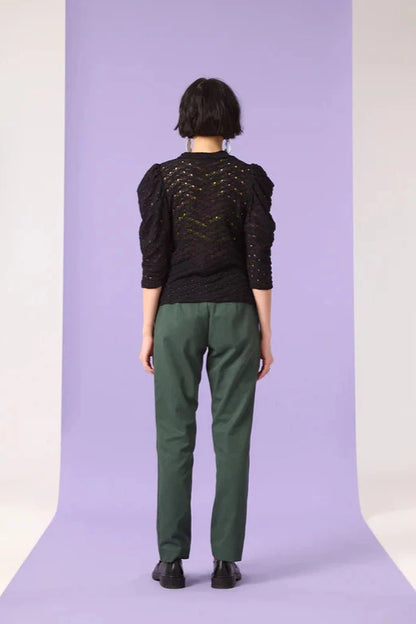 Back view of a woman wearing the Saules Pants by Cokluch in Eden, with a fly closure, an attached D-ring belt, a button and tab detail, side pockets, and an ankle length, made from OEKO-TEX certified fabric. She is wearing them with a black top and standing in front of a purple background. 